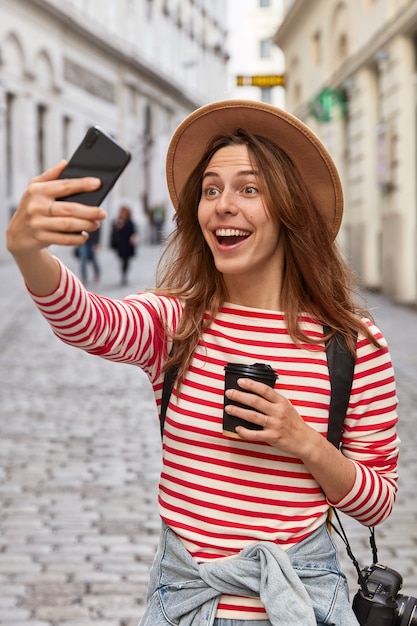 Overjoyed European female traveler in hat, makes selfie portrait outdoor, has fun during excursion in ancient town