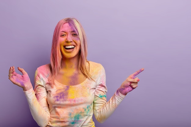 Overjoyed European woman laughs from positive impressions, shows place where Holi festival is held, has fun with colored powder, smeared with colorful dyes, smiles broadly. Celebration in India
