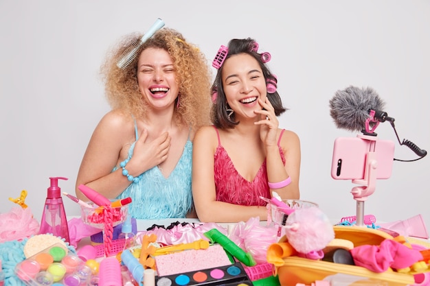 Overjoyed female beauty bloggers record video laugh happily have fun together wear dress and do hairstyle for special occasion surrounded by different cosmetic products isolated over white background