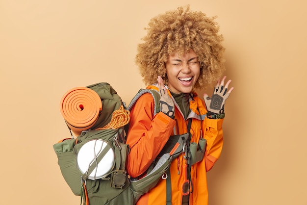 Обрадованная женщина-туристка громко восклицает, держит руки поднятыми, носит перчатки, оранжевая куртка несет тяжелый рюкзак с необходимым оборудованием, изолированным на коричневом фоне Концепция приключений в летнее время