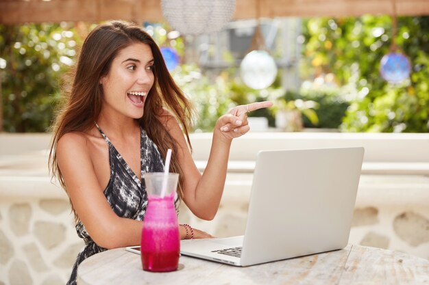 Overjoyed female with happy look and dark long hair takes break between work on laptop during lunch,