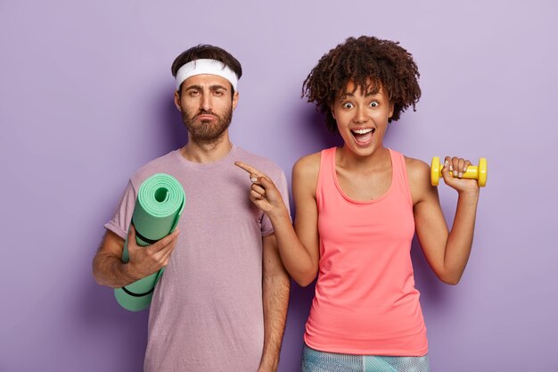 Overjoyed fit woman points at man with serious tired expression, hold sport equipment, train biceps, have yoga training with instructor. Couple in gym