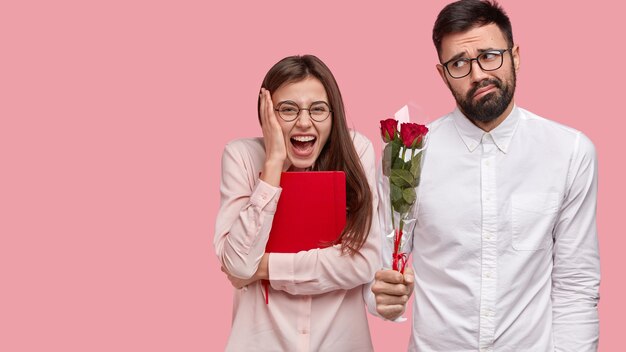 Overjoyed glad woman has first date, expresses positive emotions, awkward guy stands near with bouquet of roses