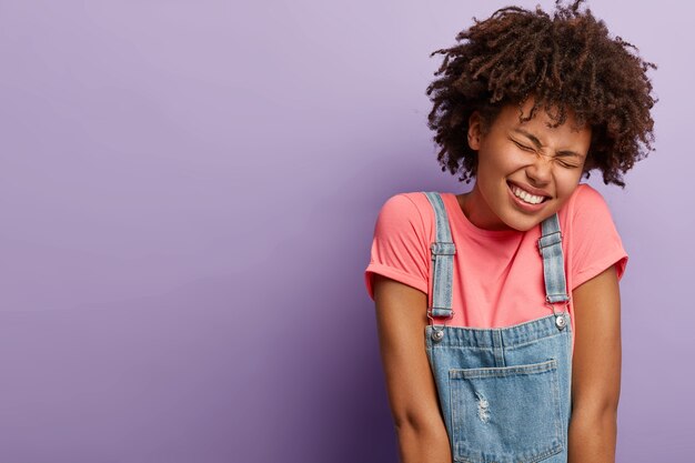 Overjoyed happy Afro American female bursts out of laughing, closes eyes, has broad smile, tilts head, hears funny joke