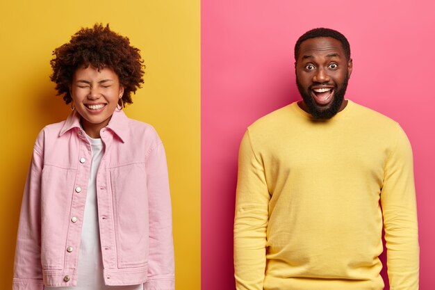 Overjoyed happy dark skinned woman and man laugh from something positive, wear casual clothes, stand against yellow and pink wall, express good emotions. Ethnicity, mood and joy concept