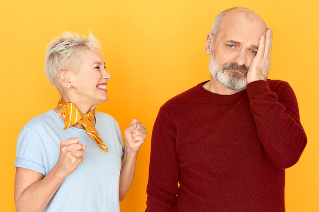 Overjoyed happy middle aged woman with blonde pixie hair clenching fists in excitement winning in lottery, her sad upset senior husband with beard holding hand on his cheek, having depressed look