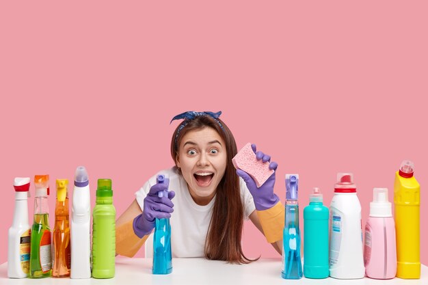 Overjoyed happy young lady keeps mouth opened, carries spray and sponge, looks in amazement, uses multipurpose detergents for scrubing