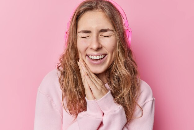 Overjoyed long haired teenage girl keeps hands pressed together laughs out gladfully closes eyes listens music via wireless stereo headphones wears casual sweatshirt isolated over pink background