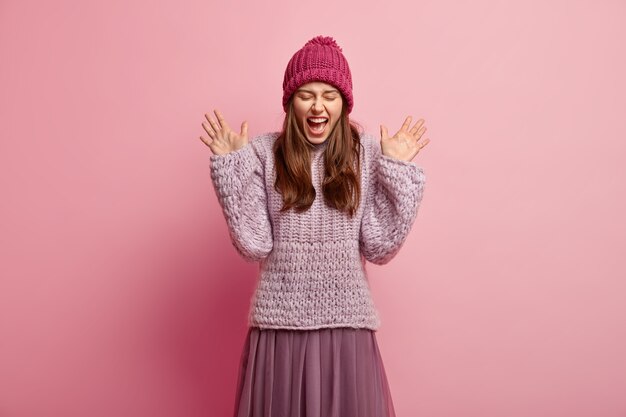 Overjoyed lovely woman with glad expression, shouts from joy and excitement, raises palms, being very emotional wears hat with pompon, knitted jumper and pleated skirt. Reaction on something pleasant