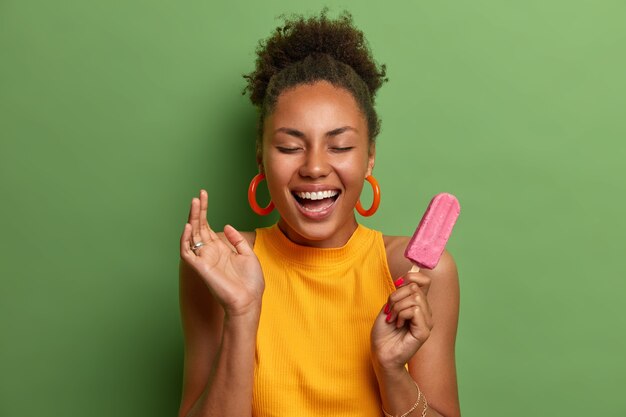 Overjoyed millennial woman with combed curly hair smiles broadly has fun and eats delicious strawberry ice cream