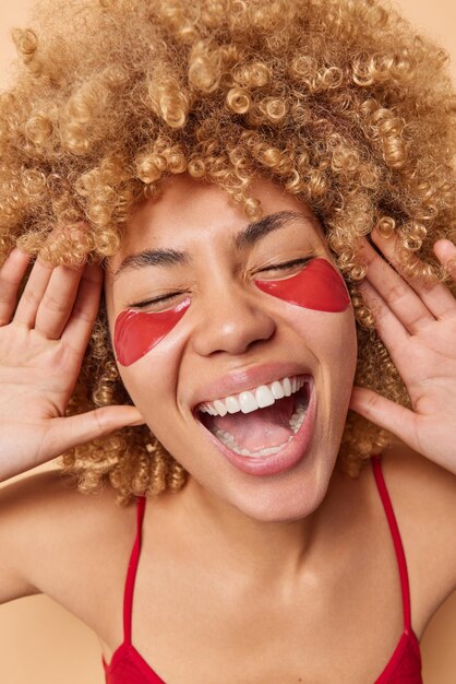 Overjoyed optimistic woman laughs out gladfully keeps mouth wide opened eyes closed applies red hydrogel patches under eyes to remove fine lines and puffiness has curly bushy hair feels glad