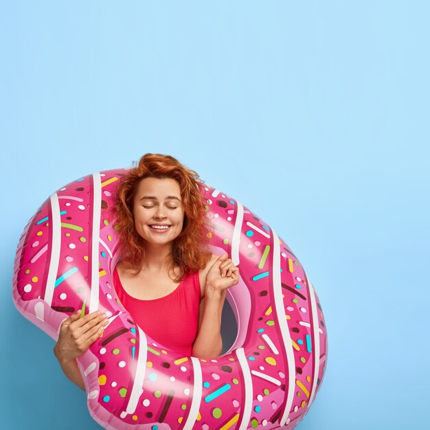Overjoyed pleased redhead woman clenches fist with satisfaction, anticipates for summer time, keeps eyes closed, stands with inflated ring, stands against blue wall with copy space. People and rest