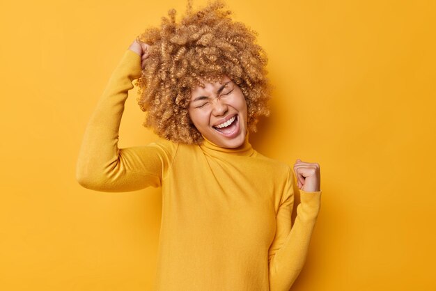 Overjoyed positive curly haired woman makes yes gesture cheers and celebrates success wears scasual jumper feels like winner isolated over vivid yellow background People and triumph concept