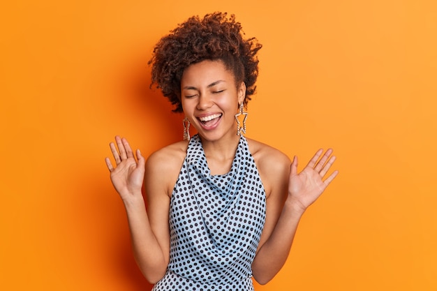 Overjoyed positive young Afro American woman has trendy hairstyle dressed in polka dot blouse smiles broadly raises palms has fun on party closes eyes poses against vivid orange studio wall