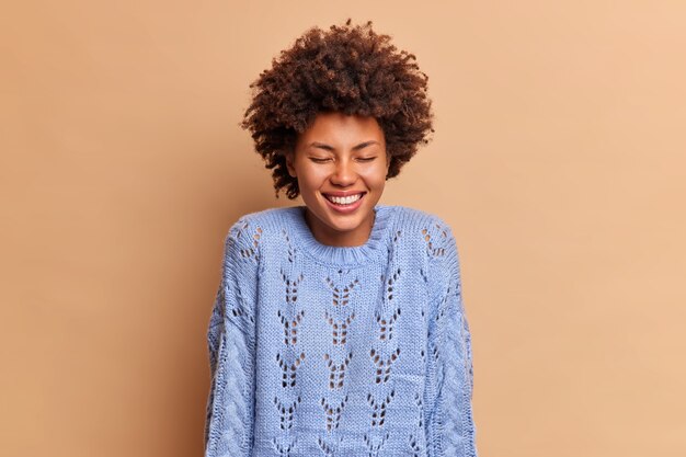 overjoyed pretty woman with curly hair smiles broadly and closes eyes in satisfaction hears something very funny dressed in casual blue jumper expresses positive emotions isolated on beige wall