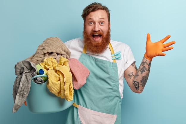 Overjoyed red haired man with foxy thick beard, raises hand, being very happy, wears casual t shirt and apron, holds basin full of laundry, has tattooed arm busy with domestic chores, glad finish work