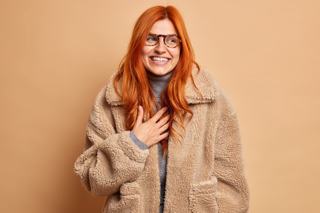 기뻐하는 빨간 머리 성인 여성이 웃고 행복한 진심을 표현하며 안경을 쓰고 따뜻한 갈색 모피 코트를 미소로 곁들여 겨울철을 즐긴다. 패션 컨셉
