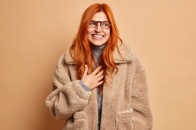 Overjoyed redhead adult woman laughs and expresses happy sincere emotions wears glasses and warm brown fur coat concentrated aside with smile enjoys winter time has upbeat mood. Fashion concept