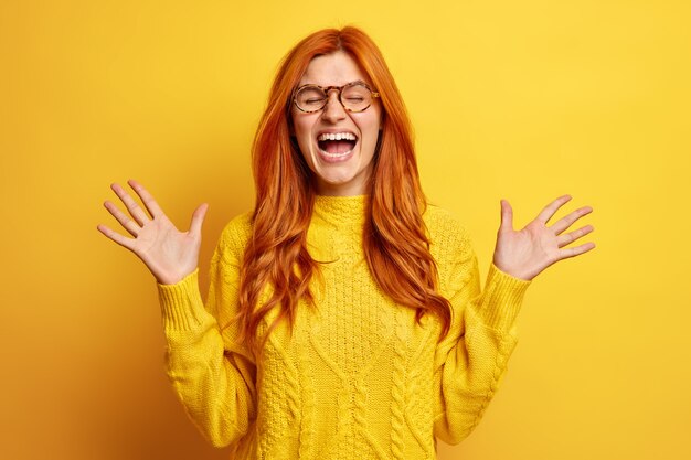 Overjoyed redhead woman laughs out loudly keeps palms raised bursts into laughter closes eyes keeps mouth widely opened wears casual jumper.