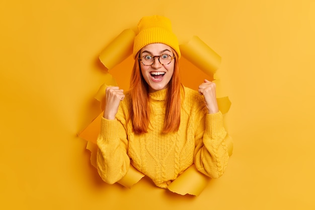 Overjoyed redhead young woman clenches fists celebrates success exclaims happily wears yellow hat and sweate