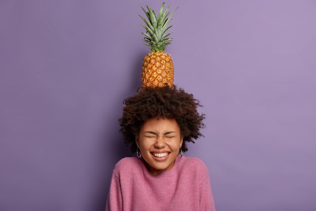 Overjoyed teenage girl poses with tasty pineapple on head, grins happily, shows white teeth, keeps eyes closed, foolishes around, has curly bushy hair
