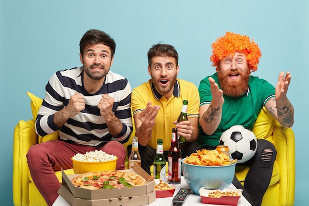 Overjoyed three best male friends being very emotional, clench fists from joy, support football team, watch game with great interest, sit on sofa, pose against blue wall. Sport supporters