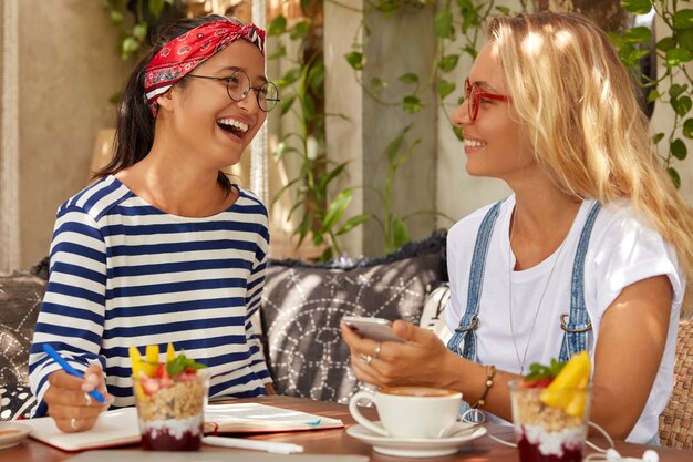 Overjoyed two women laugh happily while share opininos about planning project, communicate during coffee break, write records in organizer, eat delicious dessert, wear casual clothes and spectacles