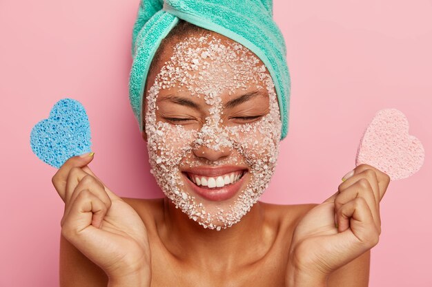 Overjoyed woman closes eyes with pleasure, smiles broadly, gets facial treatment in beauty center holds two makeup sponges poses on pink studio wall