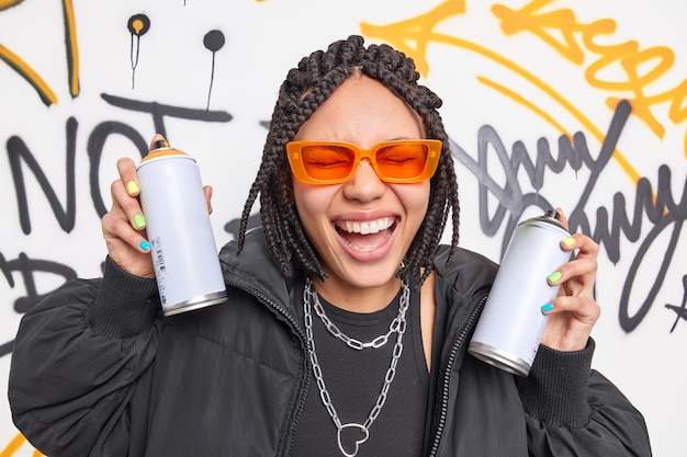 Overjoyed woman has dreadlocks feels very happy draws graffiti with aerosol spays has fun belongs to hooligan gang wears fashionable clothes laughs loudly