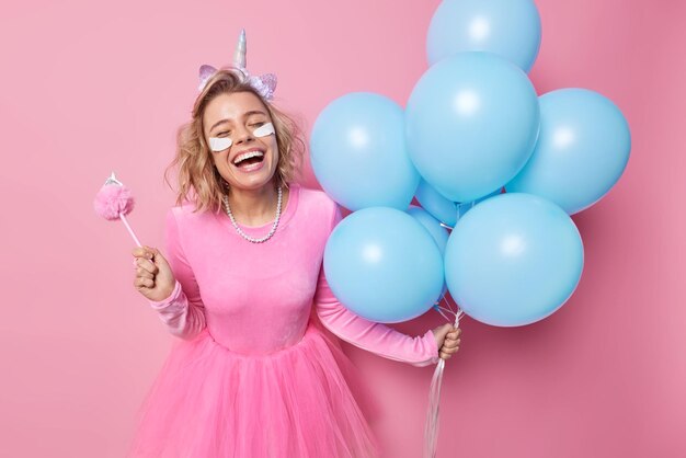 Overjoyed woman laughs happily prepaes for party holds bunch of blue inflated balloons wears dress headband and necklace applies white beauty patches under eyes isolated over pink background