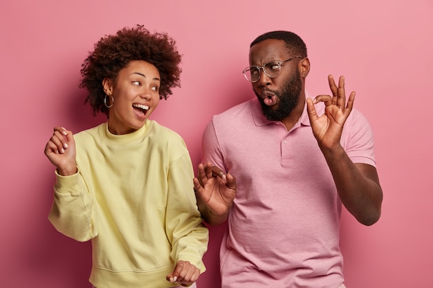 Overjoyed woman and man with dark skin have upbeat mood, dance at disco party, raise arms and move with rhythm of music, wear casual clothes, isolated on pink space. people