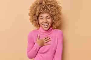 Бесплатное фото Обрадованная женщина с кудрявыми волосами, счастливо смеется, держит руку на груди, широко улыбается, носит розовую водолазку, изолированную на бежевом фоне, выражает положительные эмоции. концепция счастья