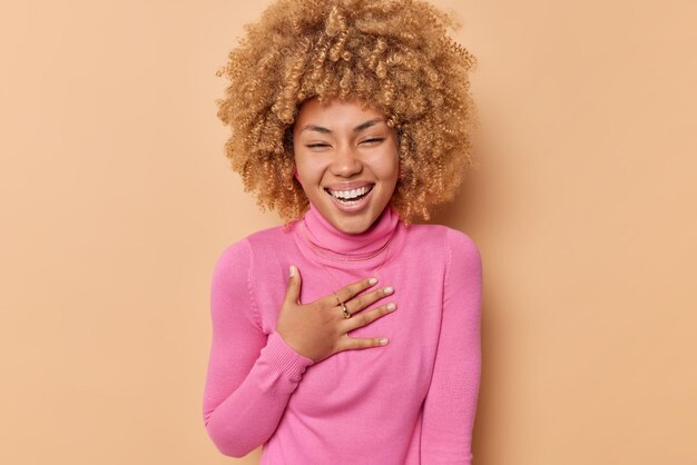 Обрадованная женщина с кудрявыми волосами, счастливо смеется, держит руку на груди, широко улыбается, носит розовую водолазку, изолированную на бежевом фоне, выражает положительные эмоции. Концепция счастья