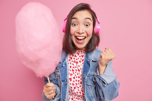 Overjoyed young Asian woman hears awesome news raises clenched fist smiles broadly celebrates triumph eats delicious sweetened candy floss listens music via wireless headphones poses indoor.