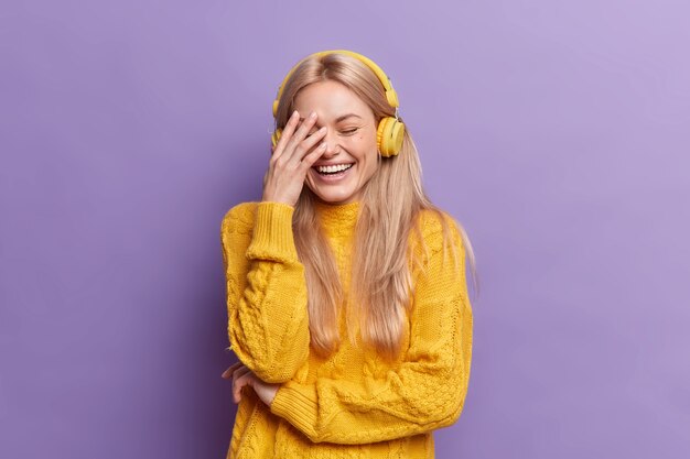 Обрадованная молодая европейская женщина со светлыми волосами громко смеется, делает лицо ладони, слушает музыку через беспроводные наушники, носит повседневный желтый свитер