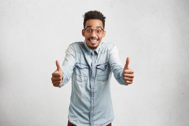 Free photo overjoyed young male with shaggy hair, mustache, raises thumbs, shows ok sign, being delightful with results of entry exams,