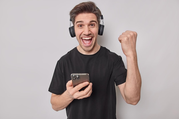 Overjoyed young man clenches fist with triumph glad to win online game uses modern technologies exclaims from joy holds mobile phone wears stereo headphones on ears dressed in black t shirt