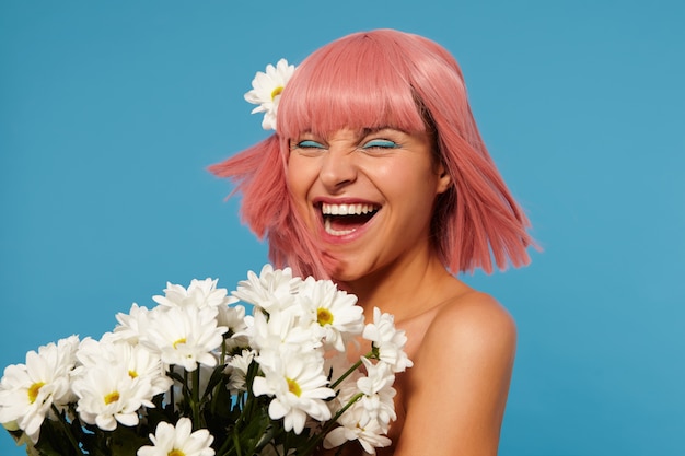 Overjoyed young pretty pink haired woman with colored makeup frowning her face while laughing happily with closed eyes, standing with white chamomiles