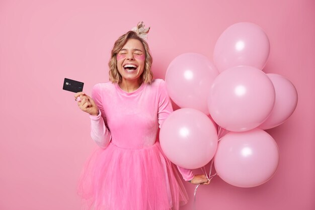 Overjoyed young pretty woman applies hydrogel patches under eyes prepares for party glad to get salary holds credit card and bunch of balloons wears festive dress isolated over pink background