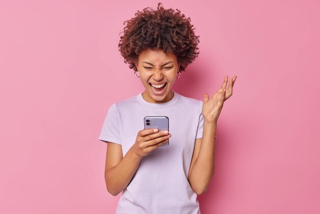 Overjoyed young woman keeps hand raised reacts on something positive concentrated at smartphone screen holds mobile phone dressed in casual t shirt isolated over pink wall