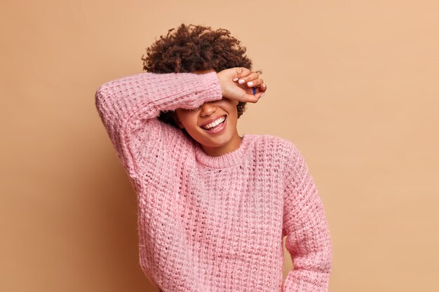 Overjoyed young woman laughs happily and smiles broadly hides eyes with arm being in good mood hears hilarious story laughs out as hears anecdote dressed in jumper isolated over brown wall