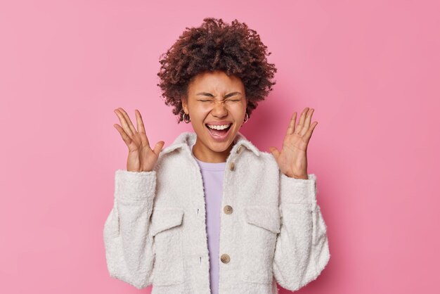 Overjoyed young woman with curly hair exclaims loudly keeps palms raised laughs happily dressed in fur jacket glad to hear awesome news isolated over pink wall