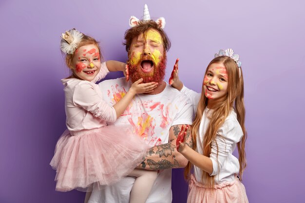 Overloaded fatigue single father with ginger beard, cries desperately, has fun with two female kids, use colourful paints, have happy expressions, stand over purple wall. Happy Fathers Day concept