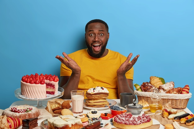 Free photo overweight addicted to sugar bearded man has puzzled expression, spreads palms, cant wait to eat desserts, surrounded by tasty cakes, cookies and milk