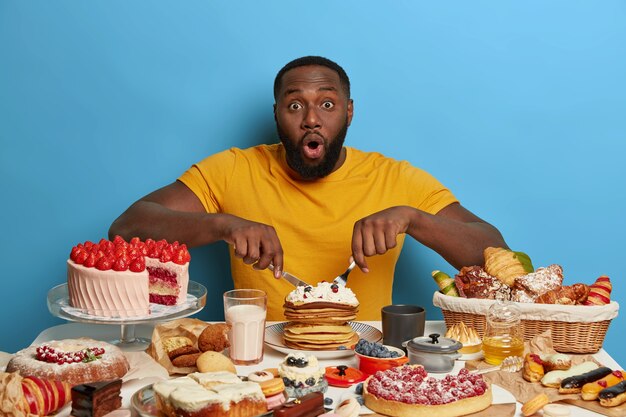 Overweight bearded young man has dark skin, surprised caught on gluttony, eats tasty creamy pancakes and other desserts, has unhealthy lifestyle