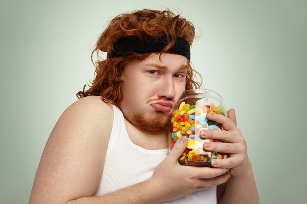 Overweight European male wearing fitness hairband and tank top after intensive cardio workout, trying hard to fight excess weight, looking unhappy, holding big jar of forbidden candies in his hands