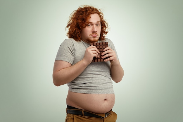 Бесплатное фото Толстый толстый мужчина с рыжими вьющимися волосами выглядит нерешительным и нерешительным, держа в руках большую плитку шоколада, в то время как ему запрещено есть сахар, и нездоровую пищу из-за строгой диеты с низким содержанием углеводов