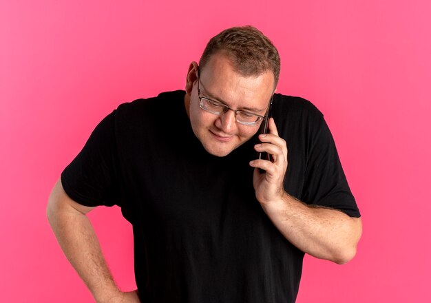 Полный мужчина в очках в черной футболке спорит во время разговора по мобильному телефону, стоя над розовой стеной