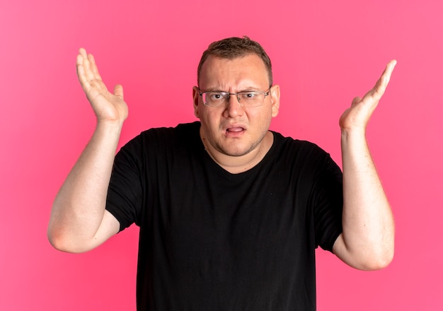 Полный мужчина в очках, одетый в черную футболку, смущен и недоволен поднятыми руками, когда спрашивает или спорит, стоит у розовой стены