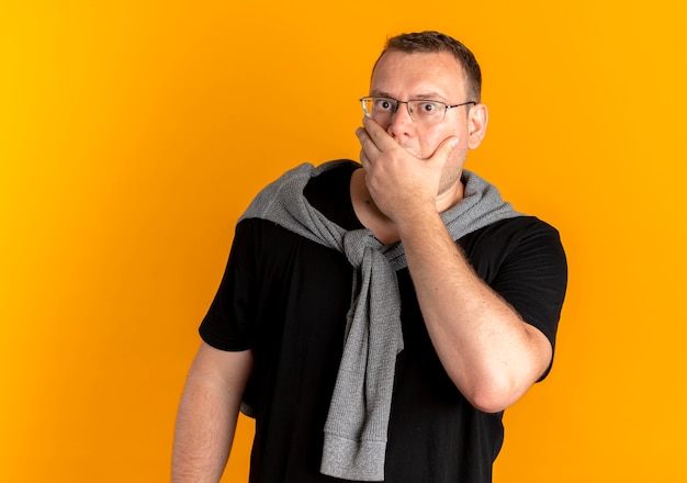 Free photo overweight man in glasses wearing black t-shirt covering mouth with hand being shocked over orange